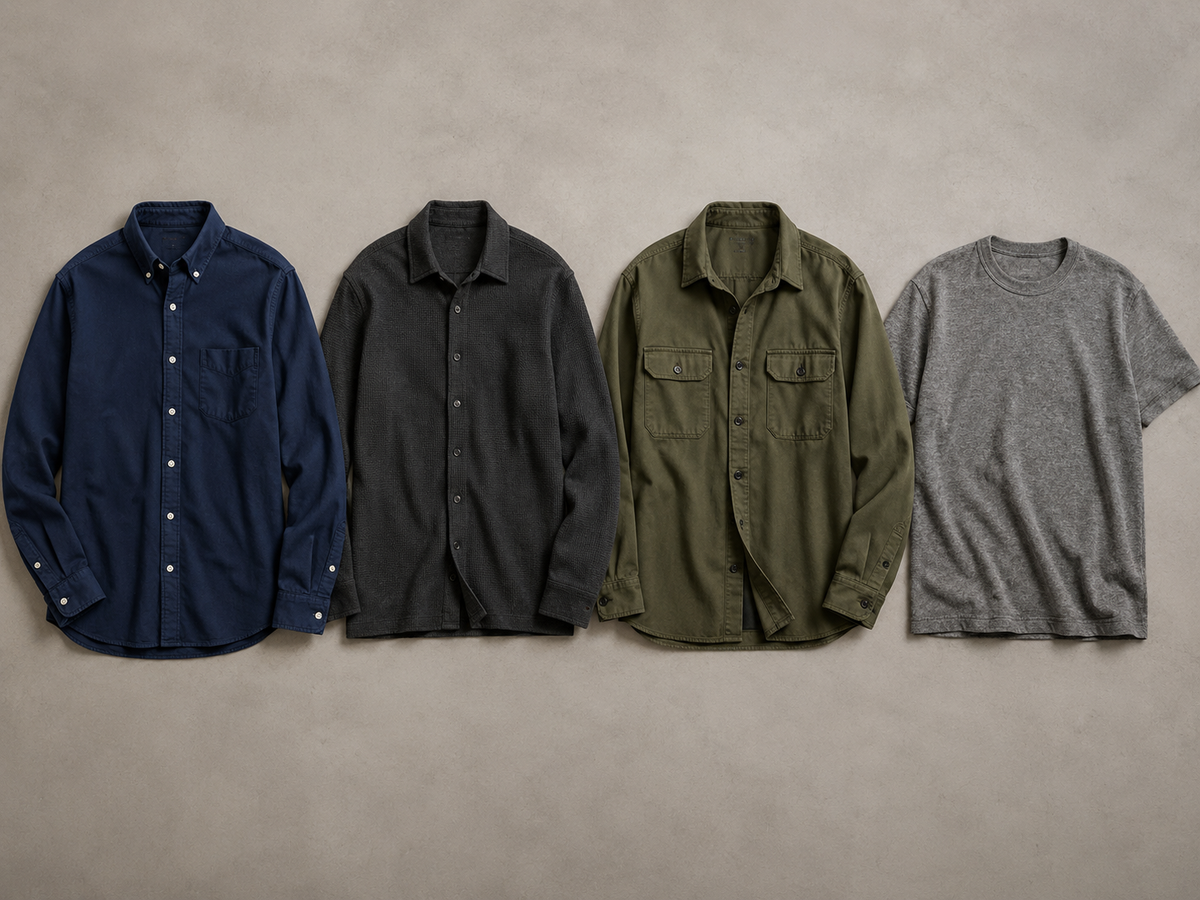 Flat-lay of four men's shirts in navy, charcoal, olive and heather grey on a warm grey surface